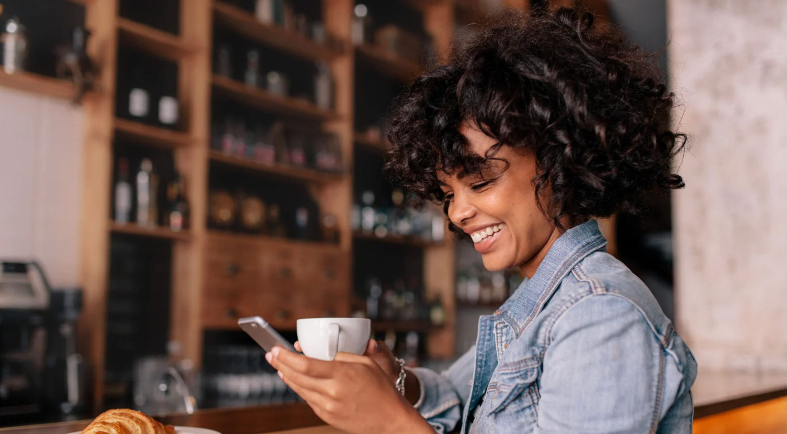 Woman smiling at smartphone at café bar counter with coffee cup